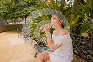 Young beautiful and happy Asian woman on park bench - lifestyle portrait of Attractive Korean girl in Summer dress drinking water