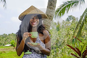 Young beautiful and happy black afro American tourist woman drinking coffee or tea visiting jungle plantation in Thailand or Vietn