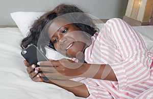 Young beautiful and happy black afro American woman in pajamas lying relaxed on bed networking with internet mobile phone online