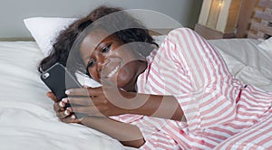 Young beautiful and happy black afro American woman in pajamas lying relaxed on bed networking with internet mobile phone online