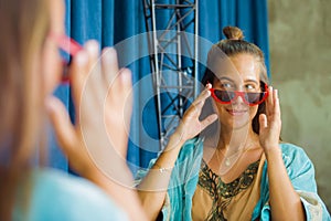 Young beautiful and happy blond woman enjoying shopping trying out clothes and sunglasses in the mirror at vintage and cool beauty