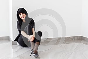 Young Beautiful happy brunette woman dressed in a black business suit sitting on a floor in office, smiling, looking at camera