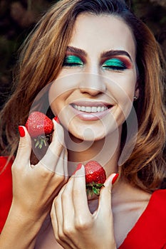 Young beautiful happy funny girl with red dress and makeup holding strawberry in summertime in the park.