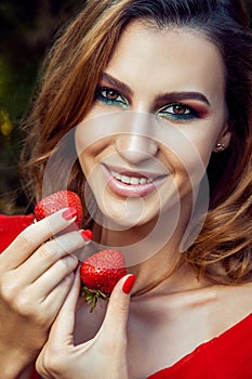 Young beautiful happy funny girl with red dress and makeup holding strawberry in summertime in the park.