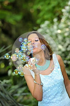 Young beautiful and happy girl blowing soap bubbles to the air on green park natural background in glamour concept