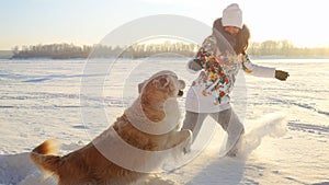 Young beautiful happy girl plays with a retriever dog in the snow in winter in sunny day during sunset time