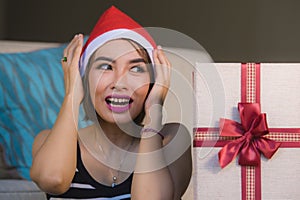 Young beautiful and happy girl in Santa hat holding Christmas present box with ribbon smiling cheerful and excited at home couch r