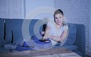 Young beautiful and happy latin woman on her 30s holding TV remote enjoying at home living room couch watching television show