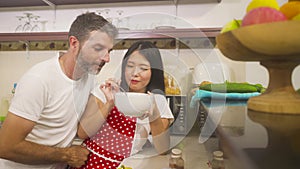 Young beautiful and happy mixed ethnicity couple in love cooking together at home kitchen the Asian Chinese woman in red apron and