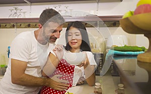 Young beautiful and happy mixed ethnicity couple in love cooking together at home kitchen the Asian Chinese woman in red apron and