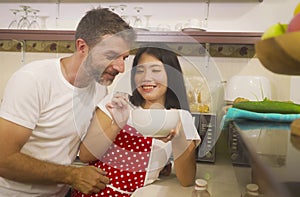 Young beautiful and happy mixed ethnicity couple in love cooking together at home kitchen the woman Asian Korean in red apron and