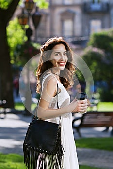 Young beautiful happy smiling lady walking on the street, holding paper cup of coffee. Model wearing stylish clothes and