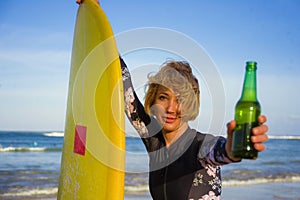 Young beautiful and happy surfer woman holding yellow surf board smiling cheerful drinking beer bottle enjoying summer holida