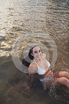 Young beautiful happy woman enjoying sunny day on the beach, sitting in the ocen water and smiling to the camera. Summer retro