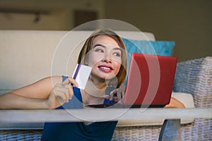 Young beautiful and happy woman at home living room holding credit card using laptop computer for banking and online shopping smil