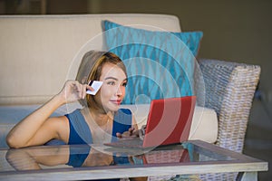 Young beautiful and happy woman at home living room holding credit card using laptop computer for banking and online shopping smil