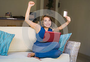 Young beautiful and happy woman at home living room holding credit card using laptop computer for banking and online shopping smil