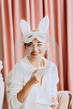 Young beautiful happy woman in a white mask of a spring Easter bunny with a cup of coffee in her hands. Carnival new year rabbit
