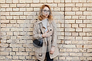 Young beautiful hipster woman in stylish glasses in a trendy spring coat in a white T-shirt with a fashionable black leather bag