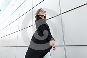 Young beautiful hipster woman in a vintage long black coat in fashionable sunglasses goes on a warm spring day
