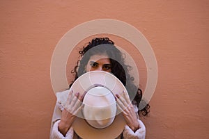 Young and beautiful Hispanic brunette woman with curly hair covers her face with her hat and exposes her eyes. The woman plays at