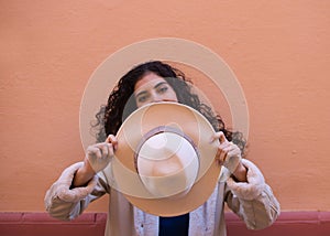 Young and beautiful Hispanic brunette woman with curly hair covers her face with her hat and exposes her eyes. The woman plays at