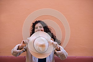 Young and beautiful Hispanic brunette woman with curly hair covers her face with her hat and exposes her eyes. The woman plays at