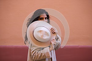 Young and beautiful Hispanic brunette woman with curly hair covers her face with her hat and exposes her eyes. The woman plays at