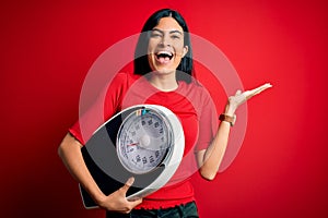 Young beautiful hispanic fitness woman holding scale for healthy weight over red background very happy and excited, winner