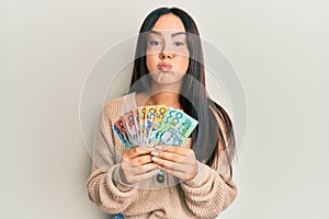 Young beautiful hispanic girl holding australian dollars puffing cheeks with funny face