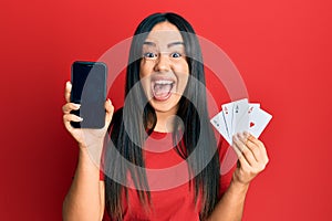 Young beautiful hispanic girl holding smartphone showing screen and poker cards celebrating crazy and amazed for success with open