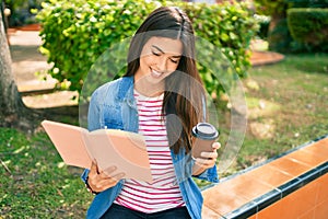 Young beautiful hispanic girl smiling happy reading book and drinking coffee sitting at the park