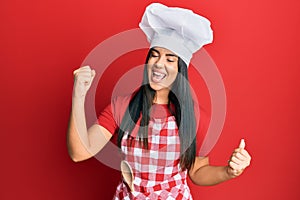 Young beautiful hispanic girl wearing baker uniform and cook hat screaming proud, celebrating victory and success very excited