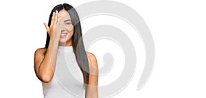 Young beautiful hispanic girl wearing casual clothes covering one eye with hand, confident smile on face and surprise emotion