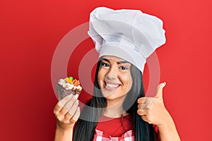 Young beautiful hispanic girl wearing chef hat holding homemade pastry smiling happy and positive, thumb up doing excellent and