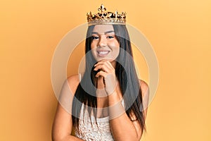 Young beautiful hispanic girl wearing queen crown smiling looking confident at the camera with crossed arms and hand on chin