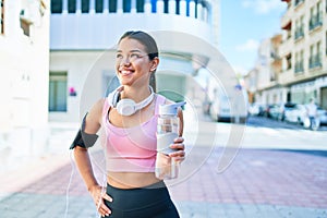 Young beautiful hispanic sport woman wearing runner outfit and headphones, drinking fresh water from the bottle