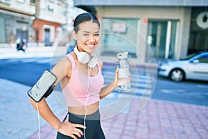 Young beautiful hispanic sport woman wearing runner outfit and headphones, drinking fresh water from the bottle