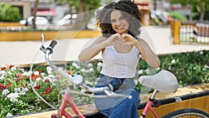 Young beautiful hispanic woman biker doing heart gesture sitting on bench at park