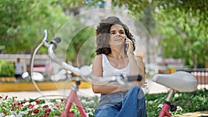 Young beautiful hispanic woman biker talking on smartphone sitting on bench at park