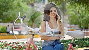 Young beautiful hispanic woman biker talking on smartphone sitting on bench at park