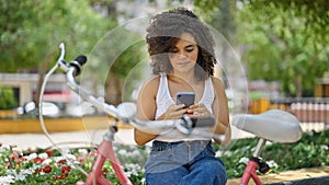 Young beautiful hispanic woman biker using smartphone sitting on bench at park