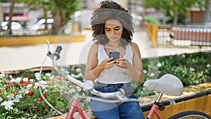 Young beautiful hispanic woman biker using smartphone sitting on bench at park