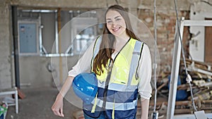 Young beautiful hispanic woman builder smiling confident holding hardhat at construction site