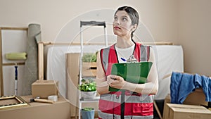 Young beautiful hispanic woman builder writing on clipboard at new home