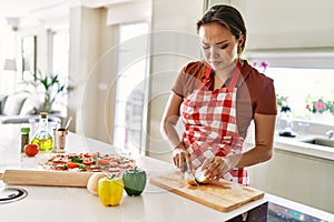 Young beautiful hispanic woman cutting onion crying at the kitchen