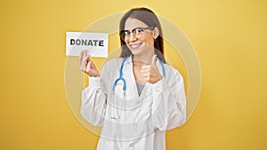 Young beautiful hispanic woman doctor doing thumbs up holding donate paper over isolated yellow background