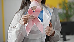 Young beautiful hispanic woman doctor holding breast cancer awareness pink ribbon and clipboard at the clinic