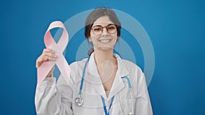 Young beautiful hispanic woman doctor holding breast cancer awareness pink ribbon over isolated blue background