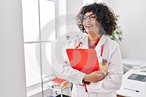 Young beautiful hispanic woman doctor smiling confident holding clipboard at clinic